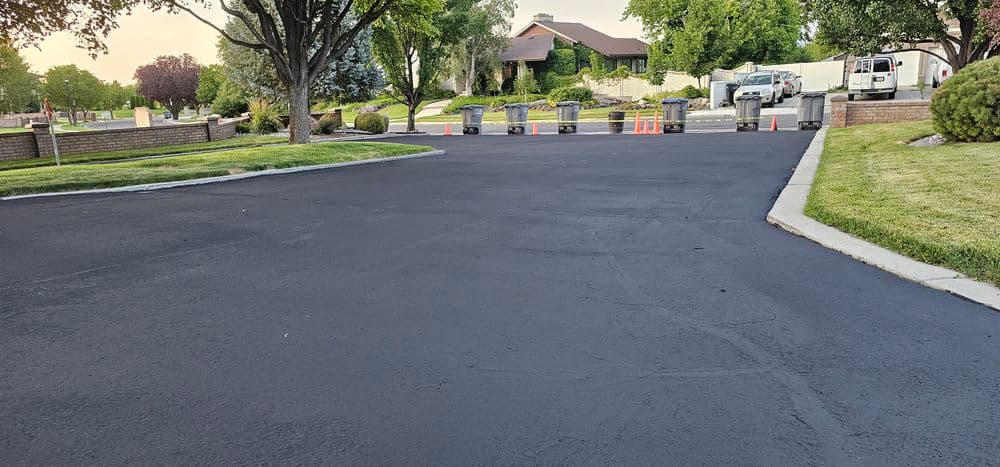 Freshly paved asphalt road with orange cones and a residential area in the background.