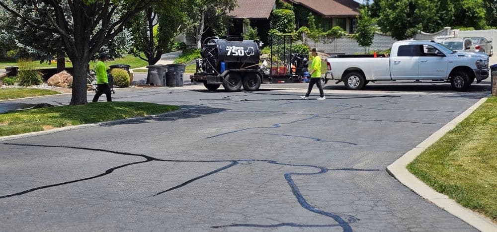 Workers repairing cracked asphalt pavement with a truck and equipment on a residential street.