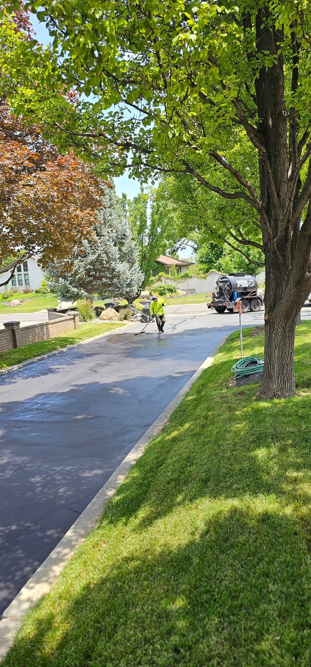 Worker cleaning a freshly asphalted driveway surrounded by trees and green grass.
