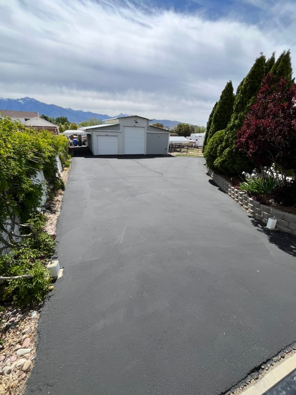 Freshly paved asphalt driveway leading to a white garage, surrounded by greenery and mountains.