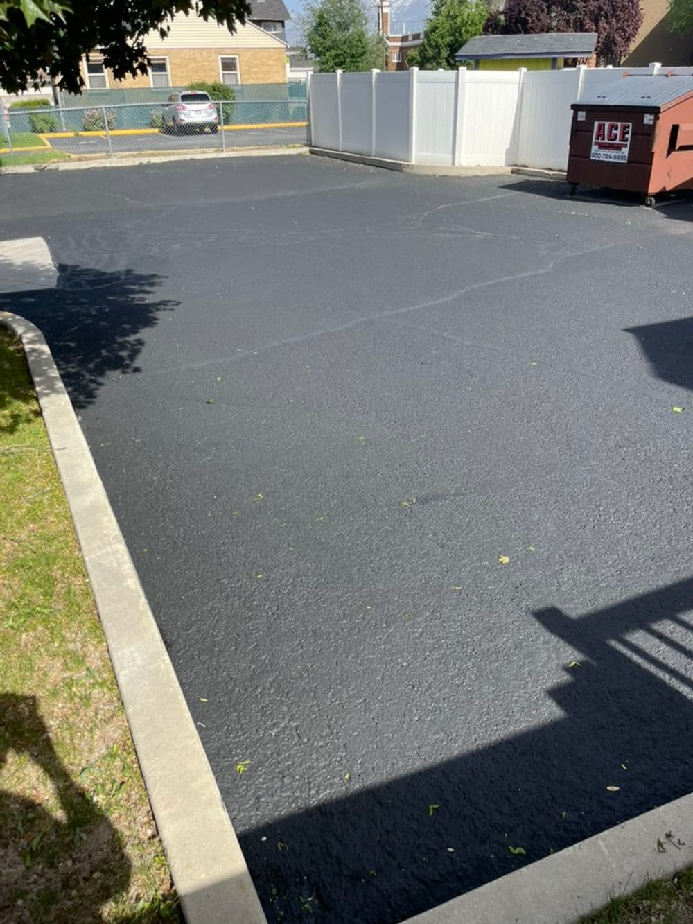 Freshly paved black asphalt parking lot with white fence and nearby dumpster.