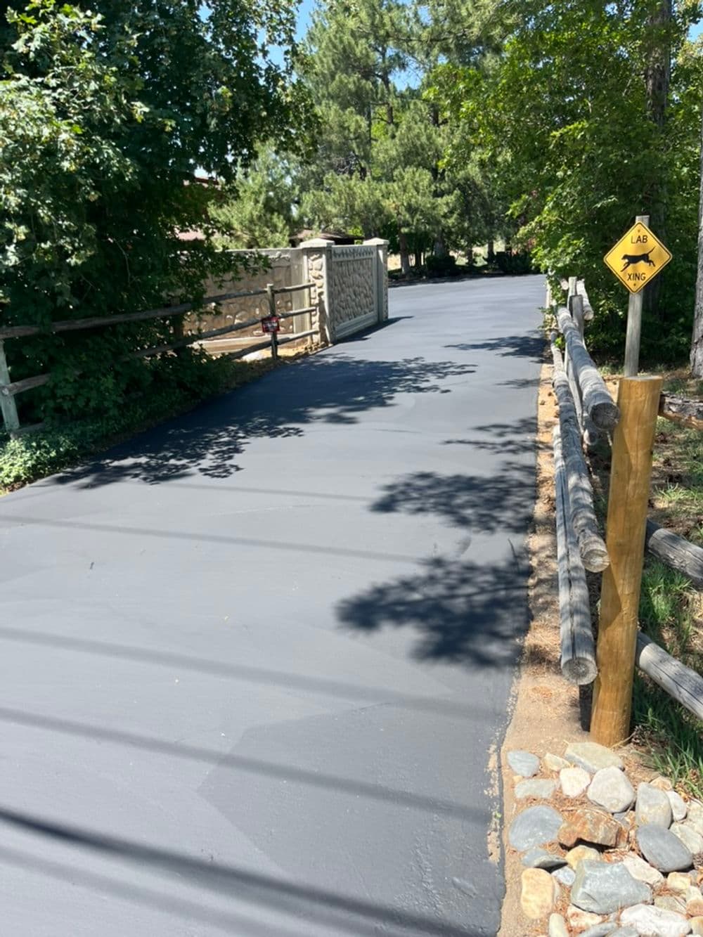 Freshly paved black asphalt road surrounded by greenery and a wooden fence. Road sign visible.