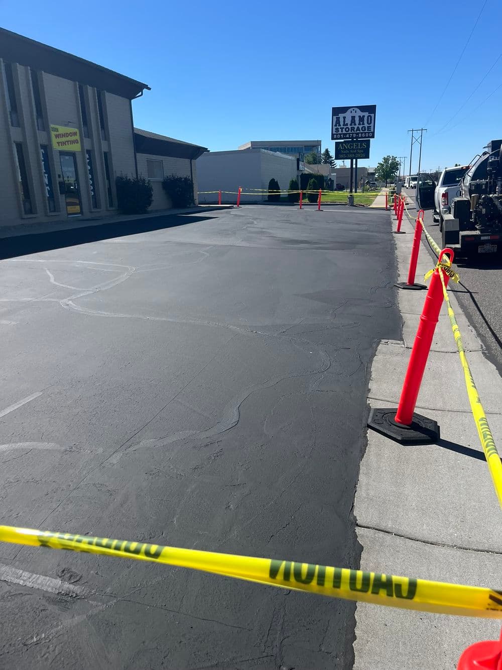 Freshly paved asphalt road with safety cones and caution tape in a commercial area.